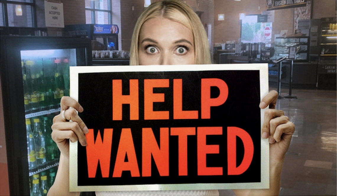 Blonde woman holding a Help Wanted sign over the bottom portion of her face.